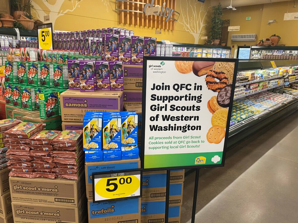 Sign reading, "Join QFC in supporting Girl Scouts of Western Washington," placed in front of a grocery store display of Girl Scout Cookies.