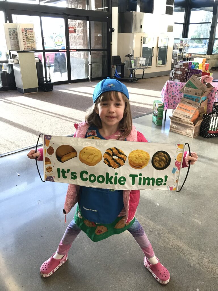 A Daisy Girl Scout holds a banner with Girl Scout Cookies and the words, "It's Cookie Time!"