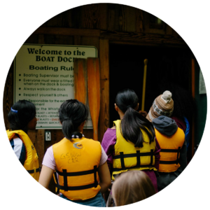 Girl Scouts stand in life jackets, looking at the boating instructions outside the boathouse.